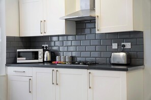 A compact kitchen with white cabinets, a black tiled backsplash, a microwave, a kettle, a toaster, and a gas stove.
