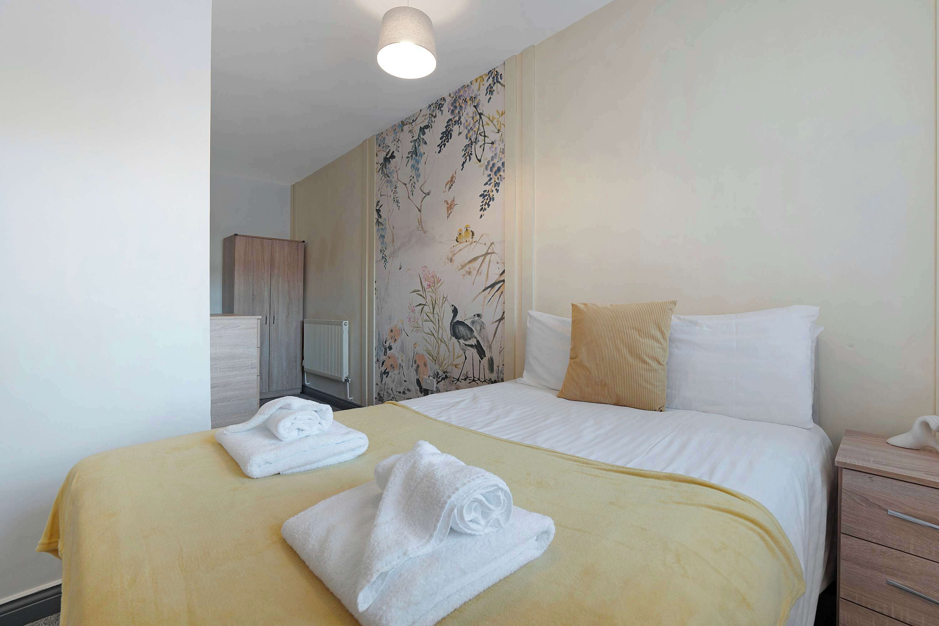 Habitación de hotel con cama, dos toallas, mesita de noche, cómoda y radiador.
