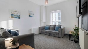 Un salon moderne avec un canapé gris, un fauteuil plus petit, une table basse et une plante en pot. Il y a une fenêtre avec des stores, un luminaire au plafond et des œuvres d’art accrochées aux murs.