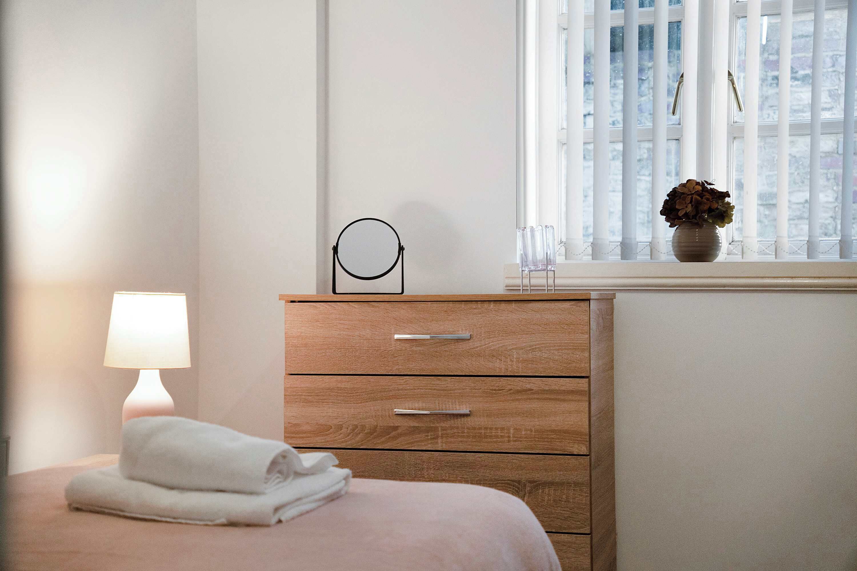 Une chambre à coucher avec un lit, une lampe de chevet, une commode à tiroirs, un miroir rond et une fenêtre donnant sur l’extérieur.