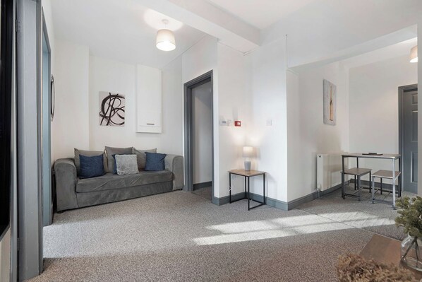 A modern living room with a grey sofa, a small side table with a lamp, and a dining area with a table and chairs.