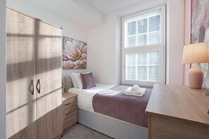 Une chambre à coucher avec un lit, une table de chevet, une commode, une fenêtre avec des stores et un tableau accroché au mur.