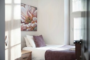 A bedroom with a bed, a nightstand, a bedside table, a window with blinds, and a wall-mounted painting of flowers.