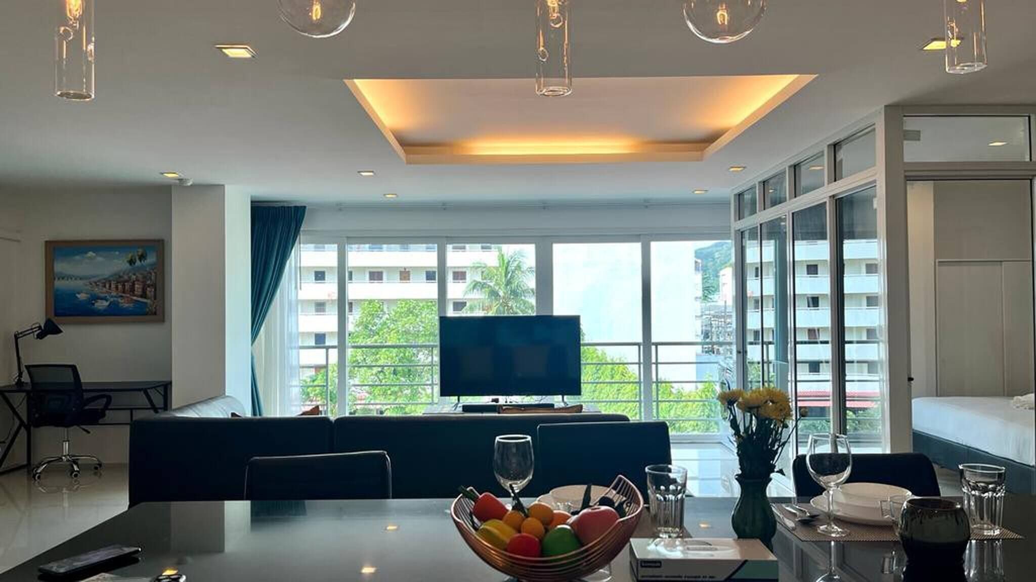 A modern hotel room with a large window, a flat-screen TV, a dining area with a table set for two, and a sofa.