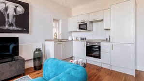 A modern kitchen with white cabinets, a microwave, and an oven. There is a large elephant painting on the wall, a blue sofa, and a television in the living area.