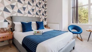 A modern hotel room with a geometric-patterned wall, a large bed with blue bedding, a bedside table with a lamp, and a view of greenery through the window.