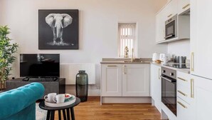 A modern kitchen with white cabinets, a microwave, and a stove. There is a large elephant painting on the wall, a TV, and a blue chair.