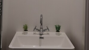 Une salle de bain avec un lavabo blanc, deux petites plantes en pot, un robinet moderne et un miroir rond doté d’un cadre noir.