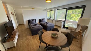 A modern room with a sofa bed, a dining table with chairs, a TV, and large windows with blinds. - Moderne Ferienwohnungen und -zimmer in Kleve (Kleve)