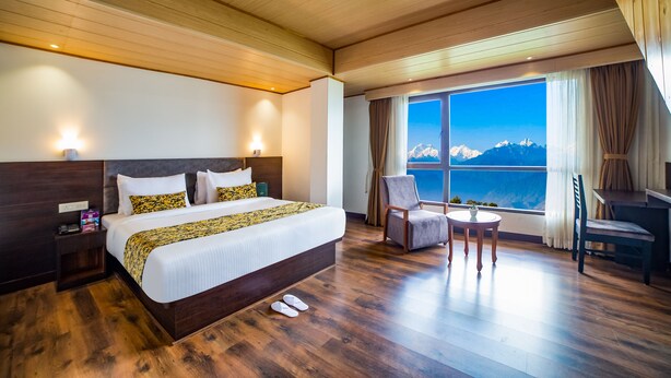 A hotel room with a large bed, a desk, a chair, and a view of mountains. - Sumitel Pelling (Gyalshing)