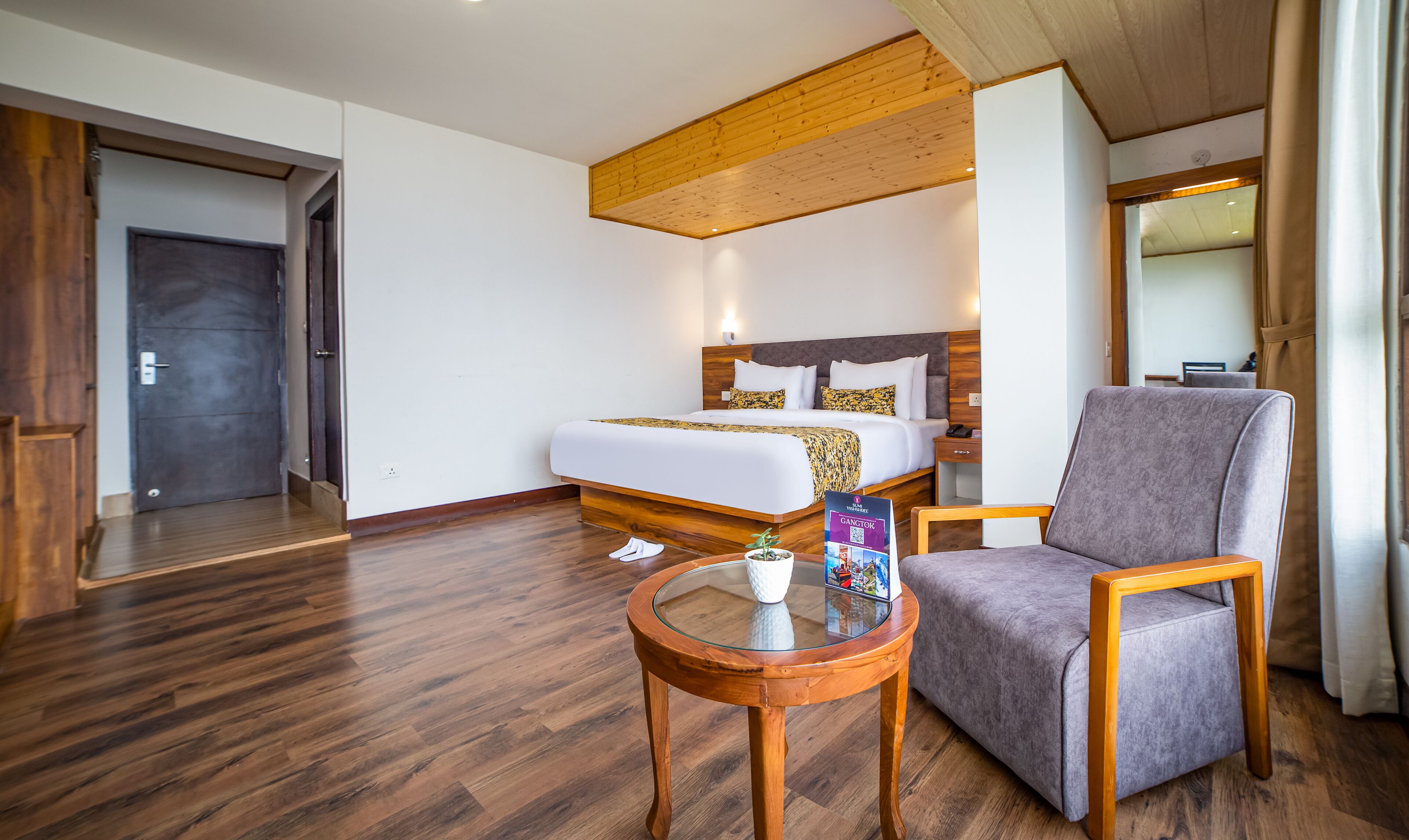 Una habitación de hotel con una cama grande, un sillón gris, una pequeña mesa de madera y suelo de madera.