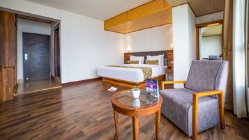 Una habitación de hotel con una cama grande, un sillón gris, una pequeña mesa de madera y suelo de madera.