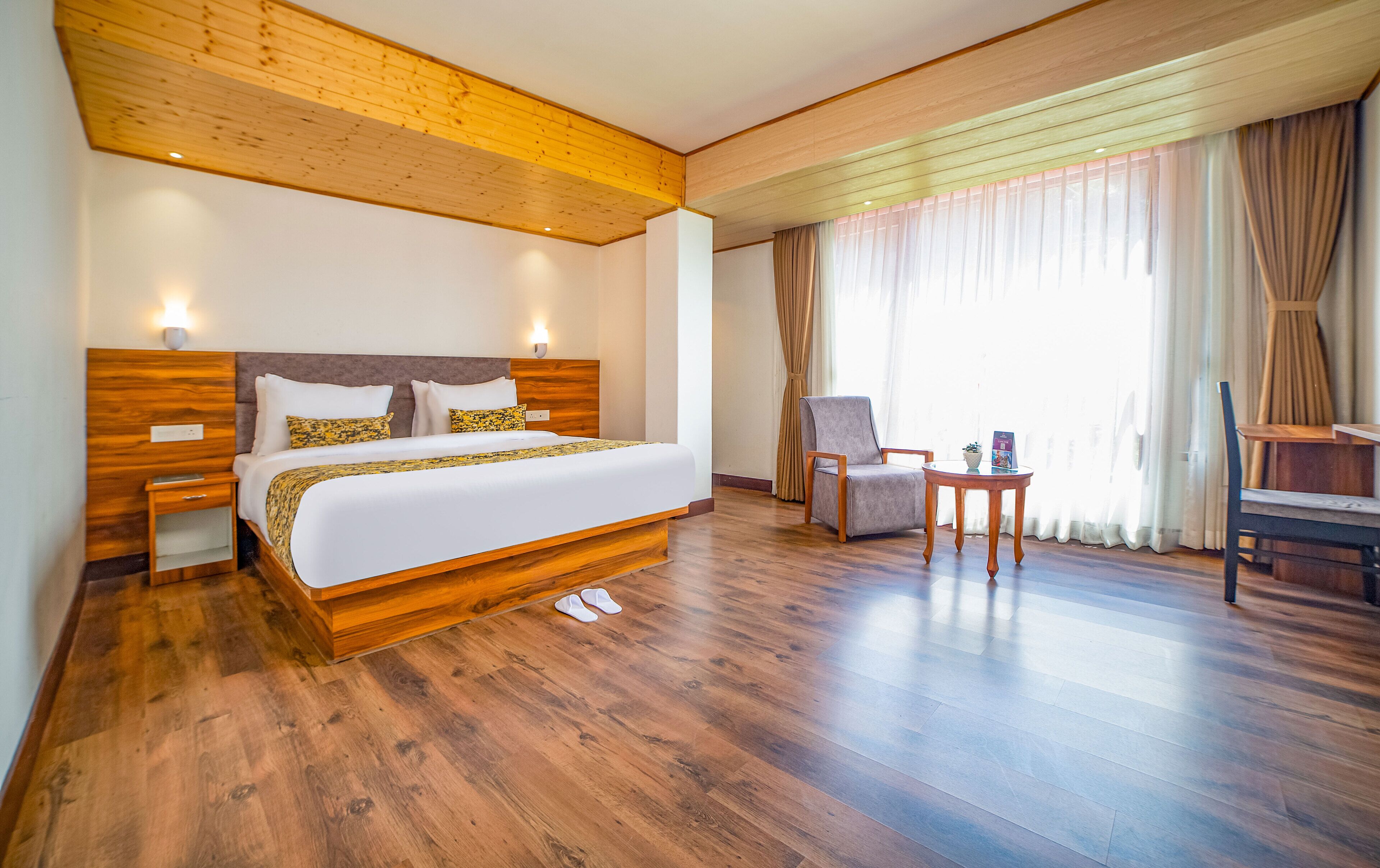 Quarto de hotel com cama de madeira, mesa de cabeceira, cadeira, mesinha e janela com cortinas.