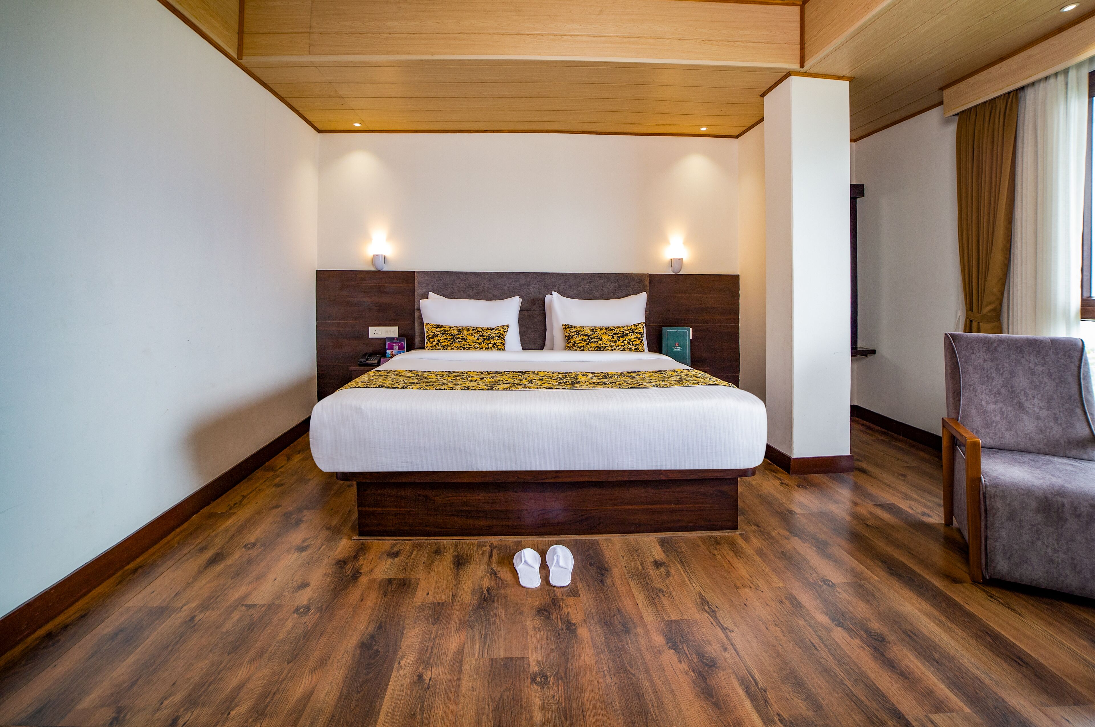 Quarto de hotel com cama grande, piso de madeira, mesa de cabeceira, cadeira e janela com cortinas.