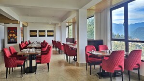 A restaurant with red upholstered chairs and tables, large windows offering a mountain view, and artwork on the walls. - Sumitel Pelling (Gyalshing)
