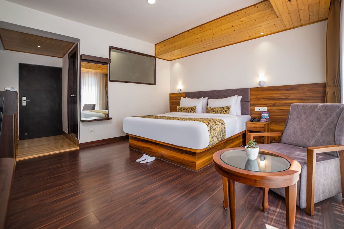 Quarto de hotel com uma cama grande, cabeceira de madeira, mesa de centro de vidro, área de estar com uma poltrona e uma televisão montada na parede.