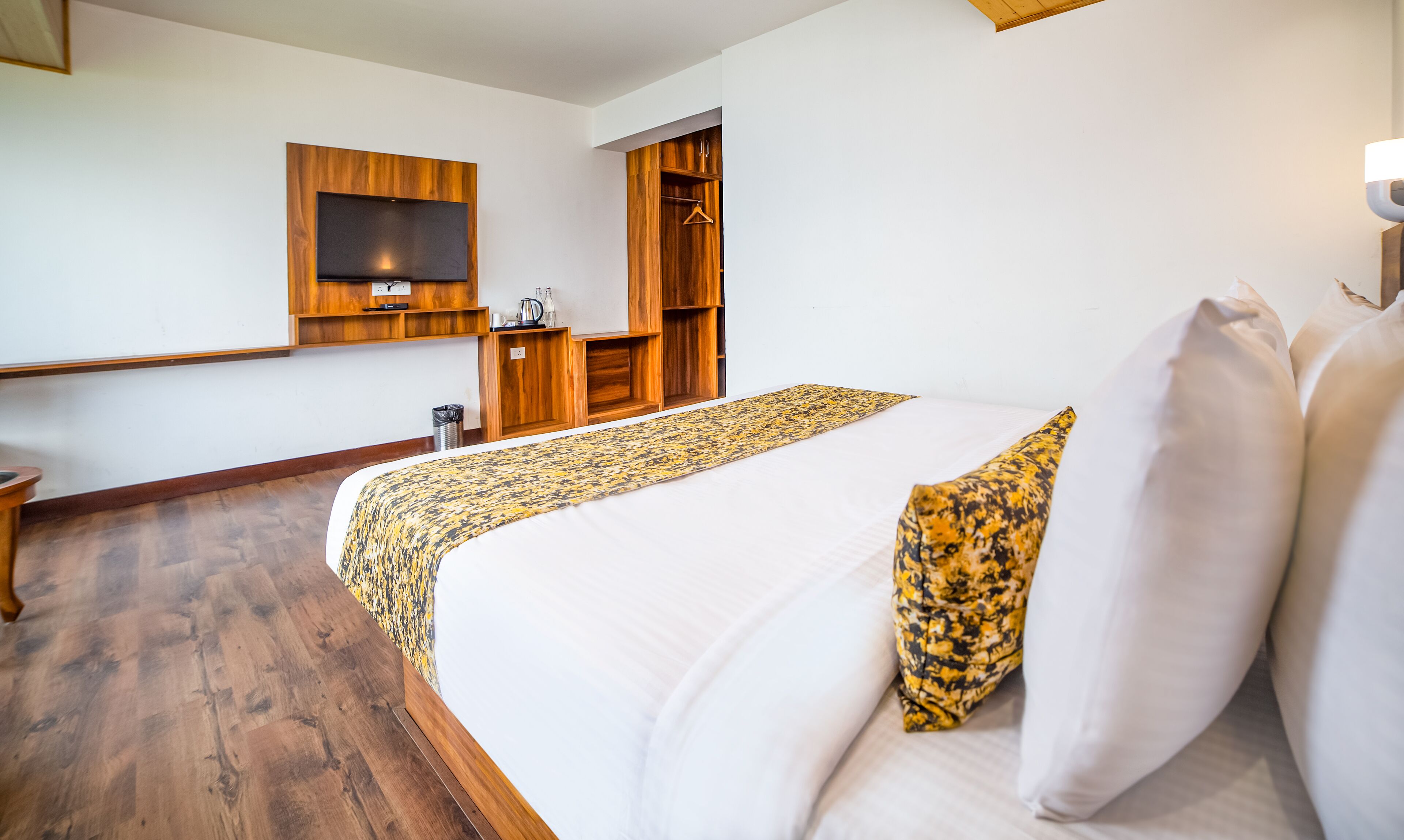 A hotel room with a large bed, a wooden wardrobe, a television mounted on the wall, and a small table with a kettle.