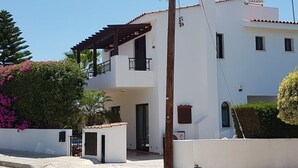 A two-story white building with a balcony, a gated entrance, and a small garden area with pink flowers. - Gracelands Emba Cypriot Village Life (Emba)