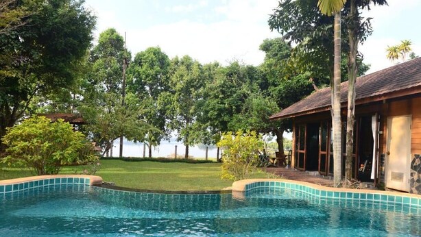 A swimming pool with clear blue water, surrounded by lush greenery and a wooden building with a tiled roof. - Viang Yonok Hotel Restaurant Sports Club (Chiang Saen)