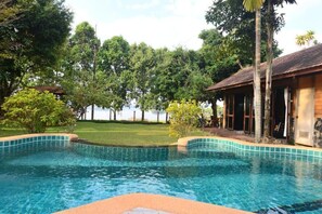 Une piscine à l’eau d’un bleu cristallin, entourée d’une végétation luxuriante et bordée d’un bâtiment en bois doté d’un toit en tuiles.