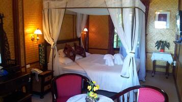A room with a four-poster bed, a small table with a vase of flowers, two chairs, a nightstand with a lamp, and a wall-mounted shelf with a plant.