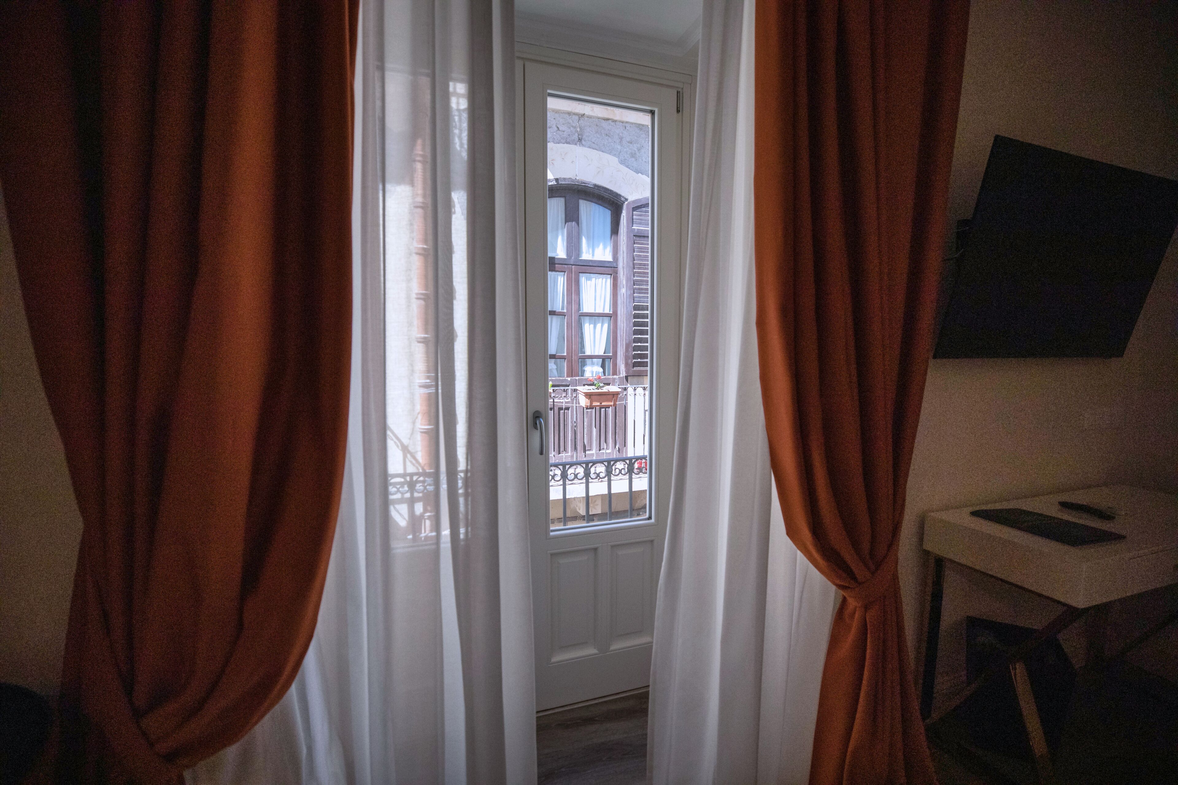 Een deuropening met witte gordijnen, een klein tafeltje met een boek, en uitzicht op een balkon met een hekwerk.