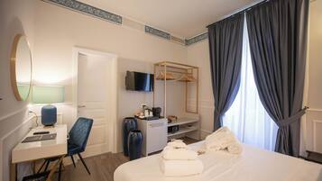 Quarto de hotel com cama, escrivaninha com cadeira, televisão, guarda-roupa e janela com cortinas.