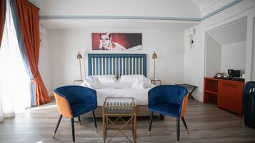 A hotel room with a bed, two blue chairs, a small table, a painting on the wall, and a desk with a lamp.