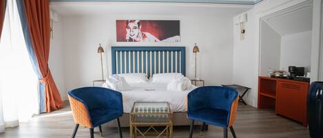 Une chambre d’hôtel avec un lit, deux chaises bleues, une petite table, un tableau au mur et un bureau avec une lampe.
