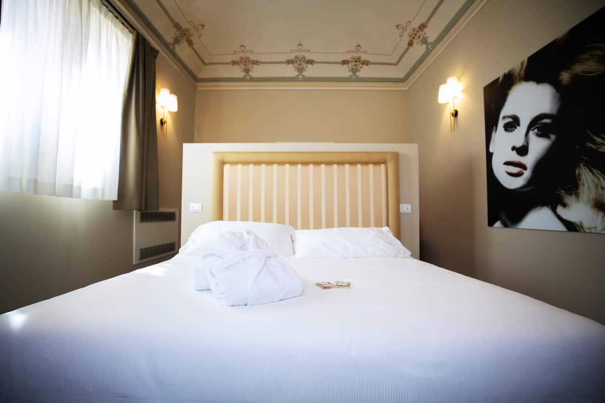 Et hotelværelse med en stor seng, hvide sengetøj, en sengegavl, to væglamper og et stort portræt på væggen.