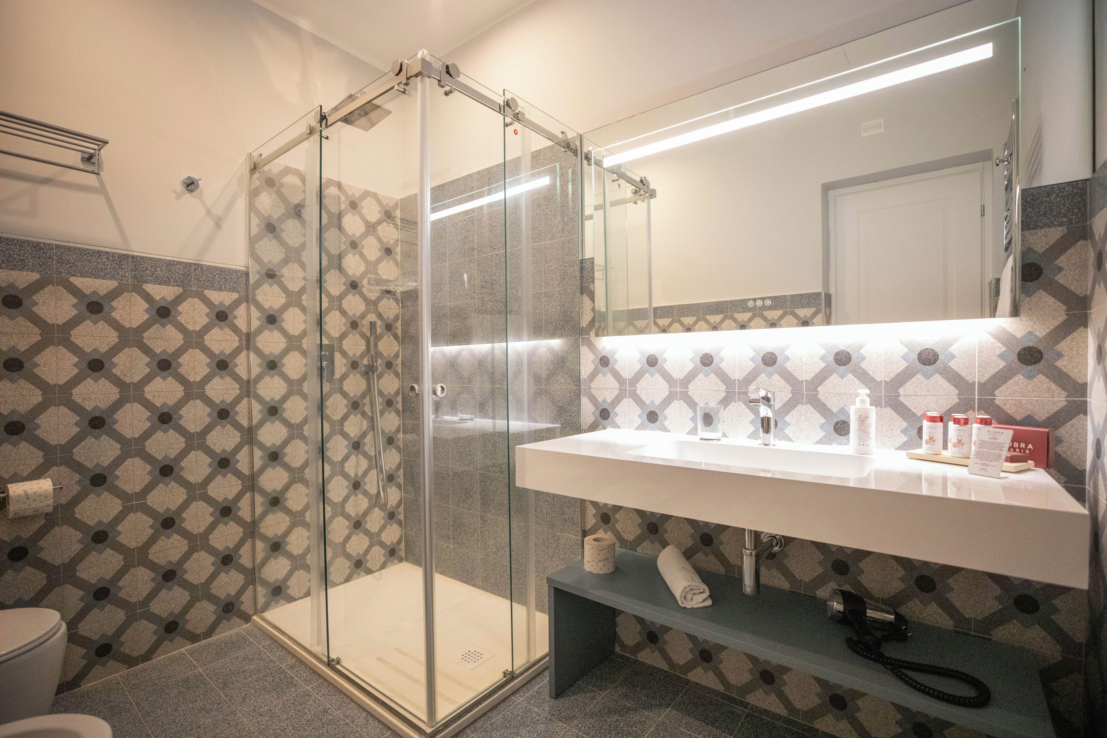 A modern bathroom with a glass-enclosed shower, a white sink with toiletries, and patterned tile walls.