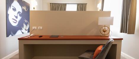 A hotel room with a desk, chair, and a large framed artwork on the wall.
