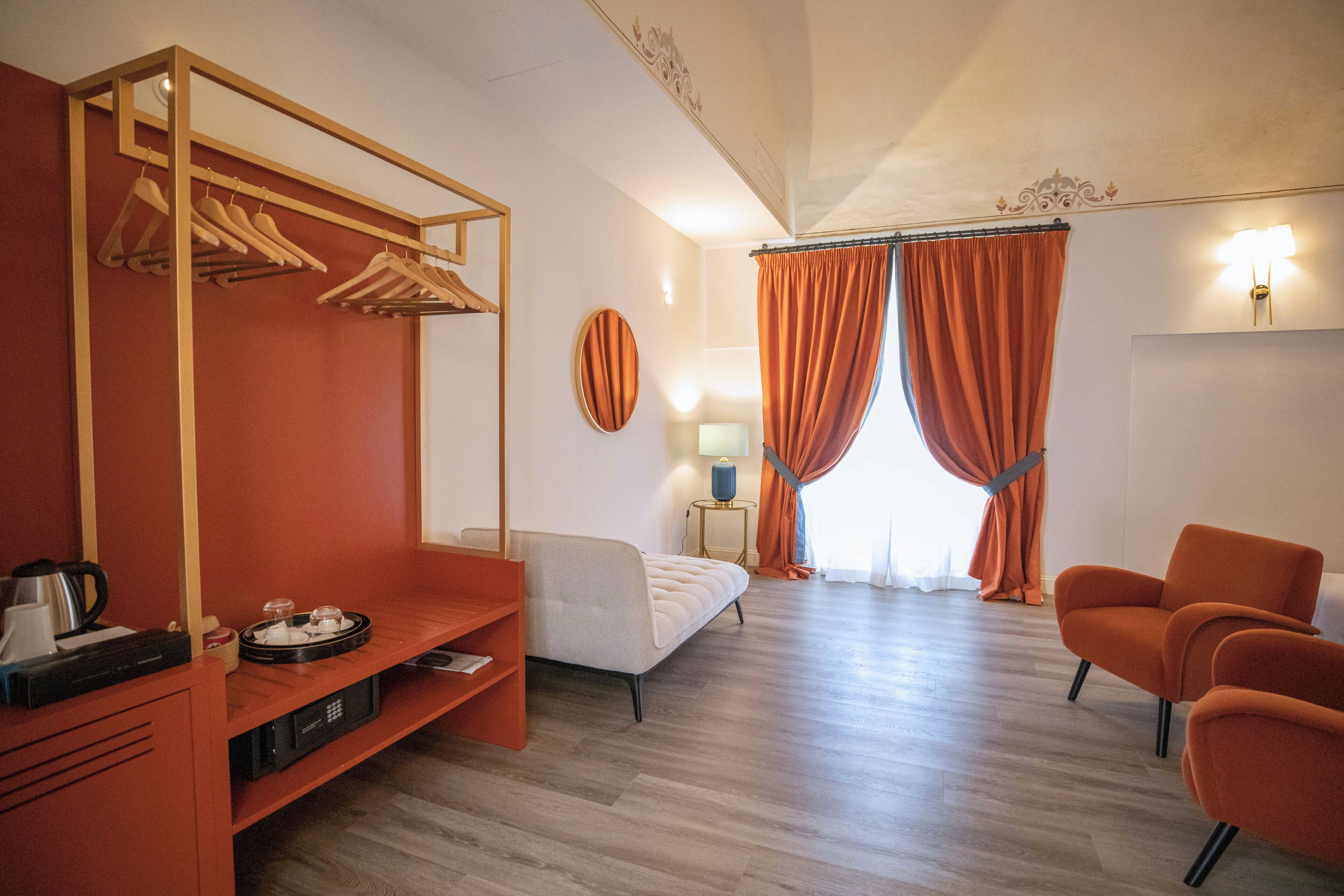Habitación de hotel con cama, zona de estar con una silla, estante de madera con una radio y una tetera, perchero y ventana con cortinas.