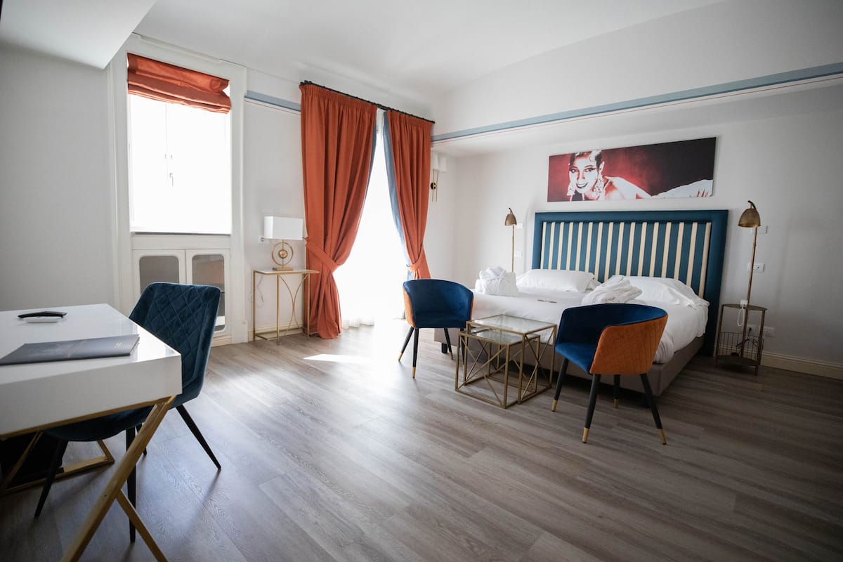 Et hotelværelse med seng, skrivebord, to stole, lampe og et vindue med gardiner.