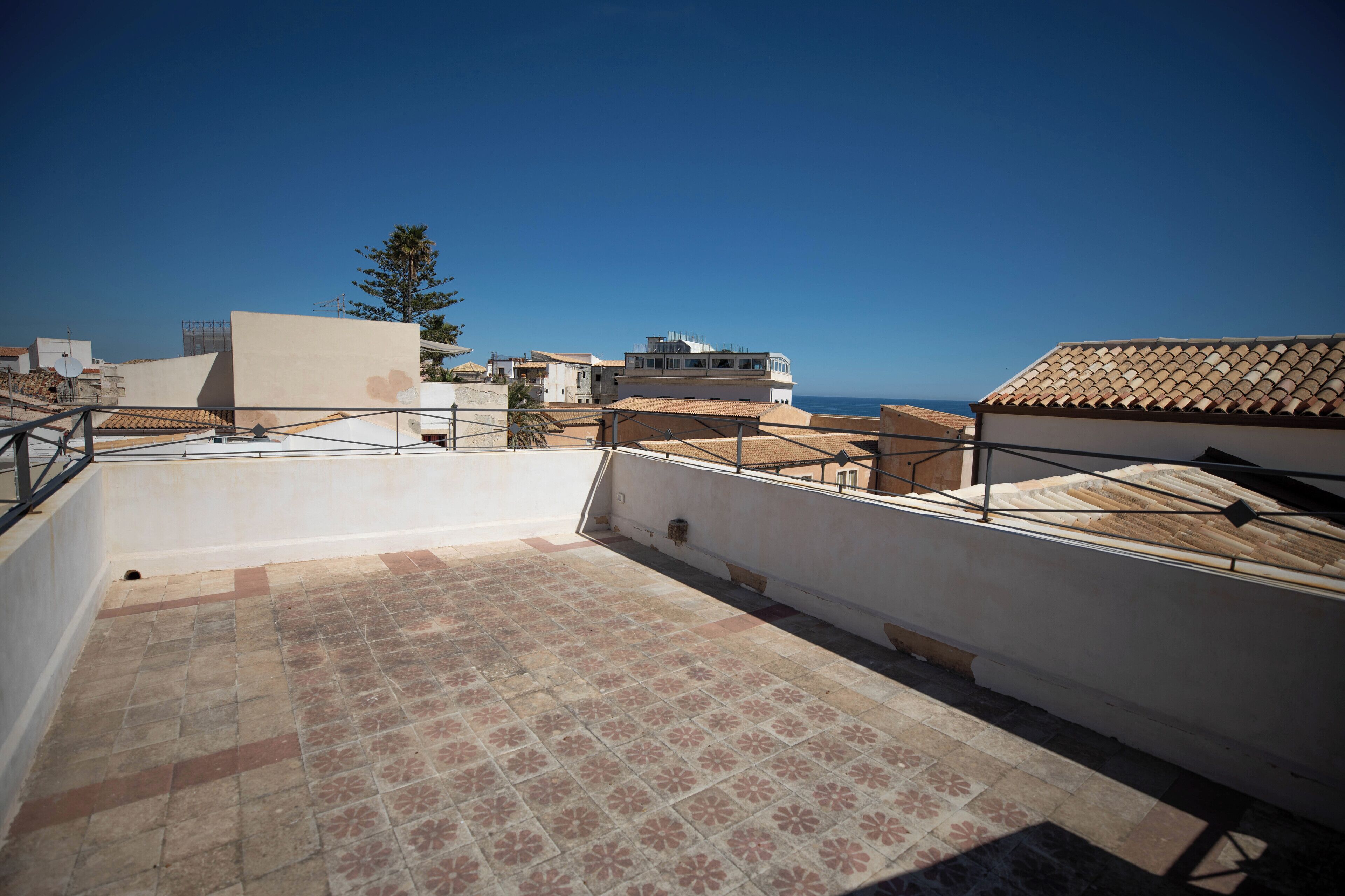 A rooftop terrace with tiled flooring, a railing, and a view of the sea and buildings.
