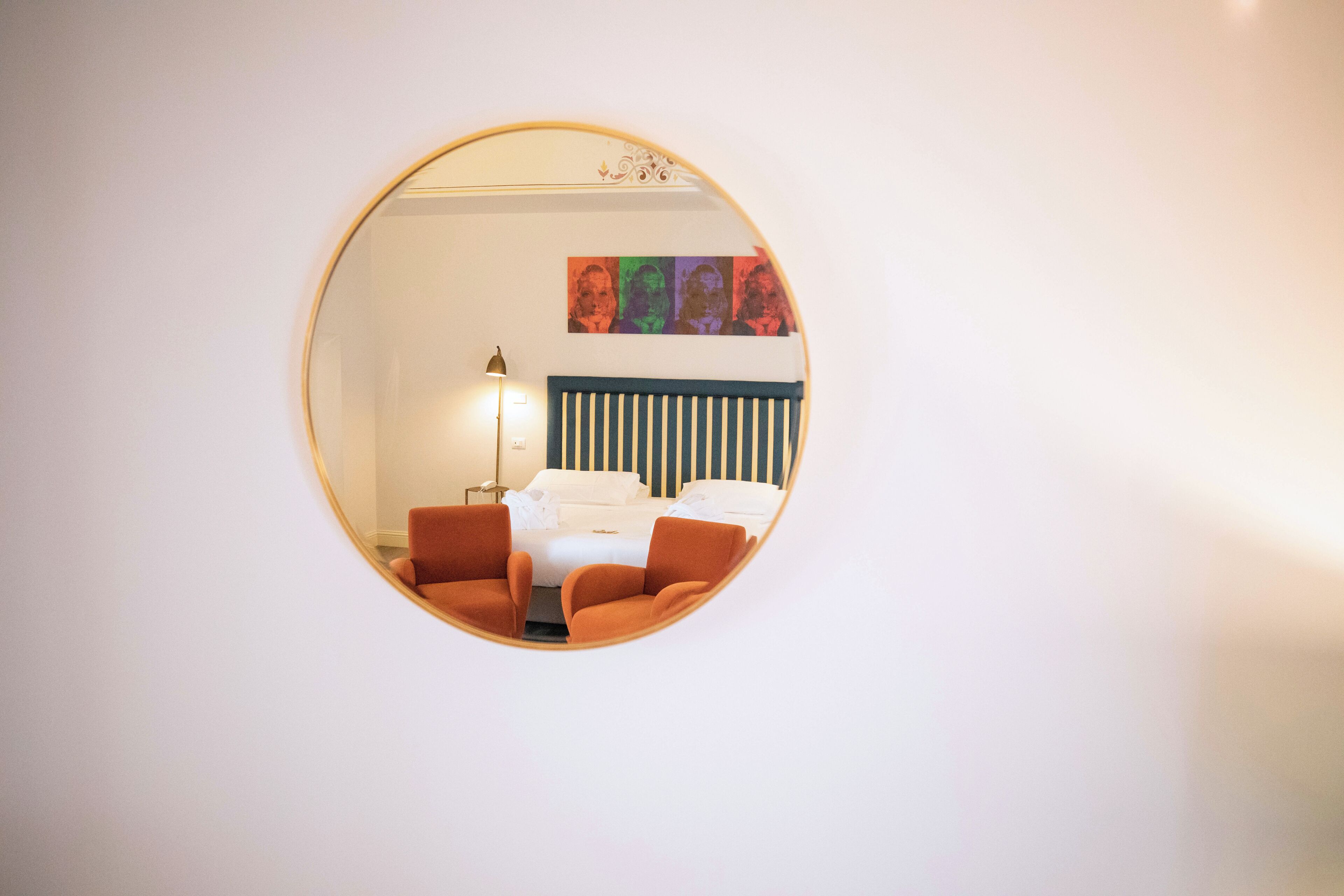 A round mirror reflecting a room with a bed, two orange chairs, a floor lamp, and a wall with a striped headboard.