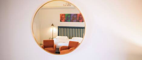 A round mirror reflecting a room with a bed, two orange chairs, a floor lamp, and a wall with a striped headboard.