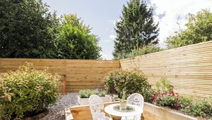 Une terrasse avec une table ronde et des chaises blanches, entourée de jardinières en bois surélevées et d’une clôture en bois.