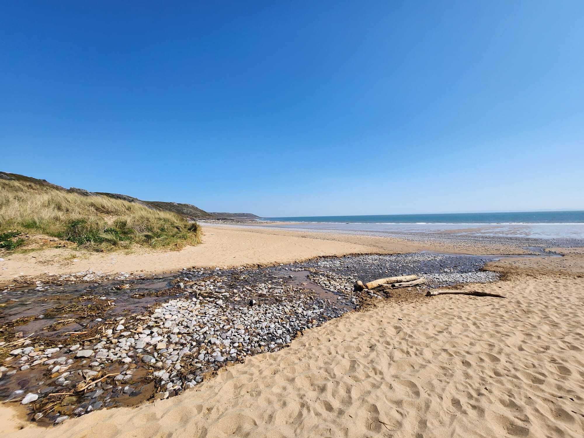 Una spiaggia con ciottoli e sabbia, un cielo sereno e una collina verdeggiante sullo sfondo.