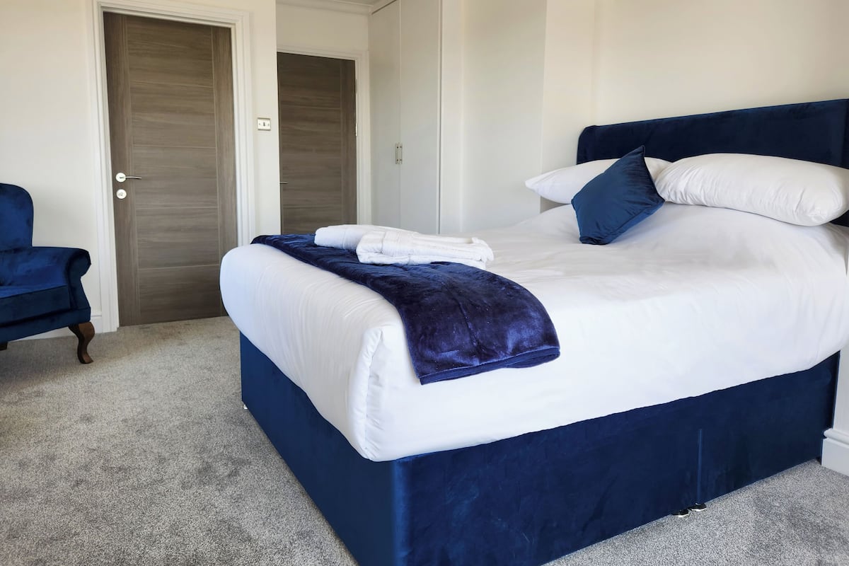 Un lit bien fait, avec une literie blanche et bleue, une tête de lit bleu foncé et une chaise bleue, dans une pièce dont le sol est recouvert d’une moquette et qui possède une porte en bois.