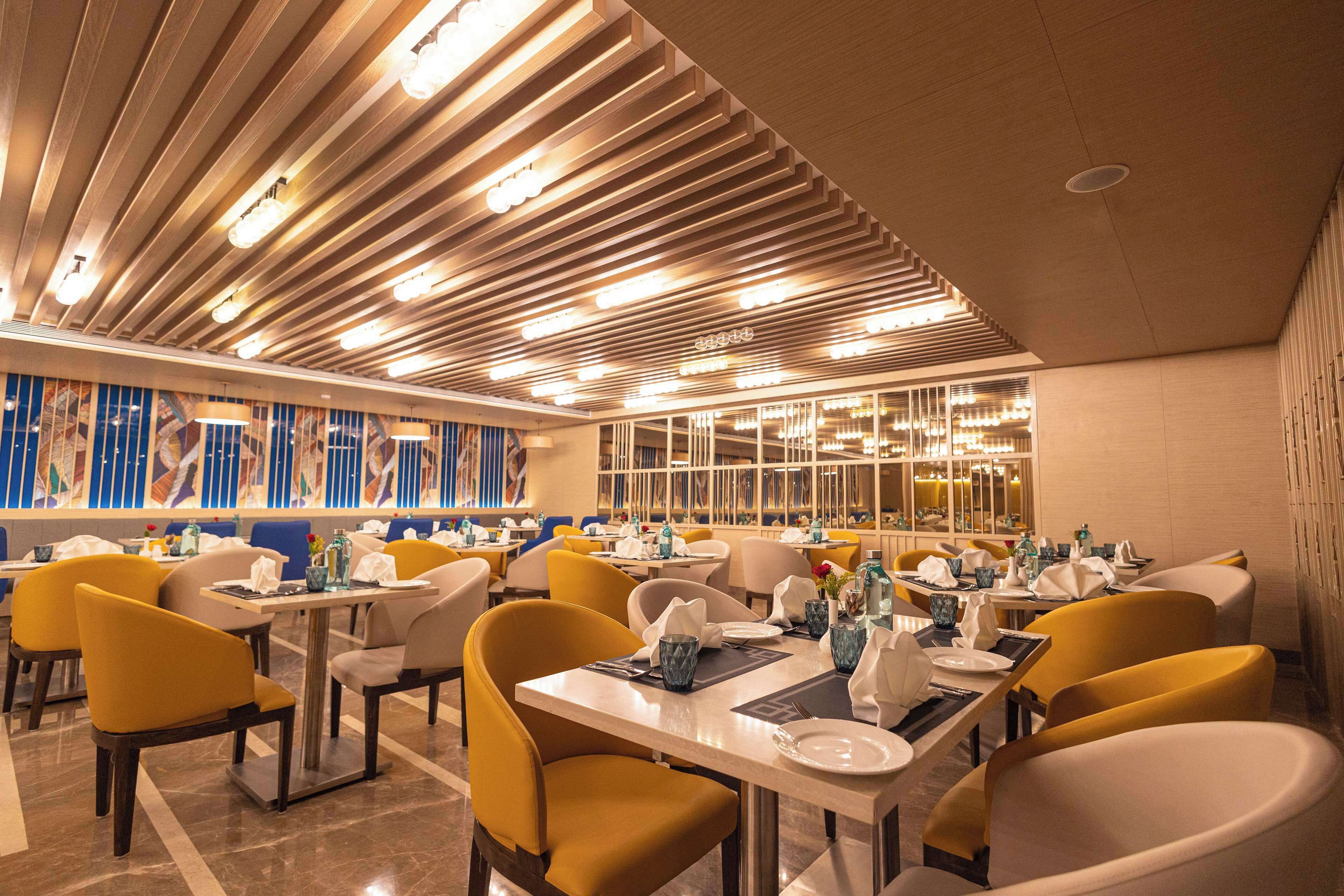 A modern restaurant with yellow chairs, tables set with plates and glasses, and a decorative ceiling with linear lights.