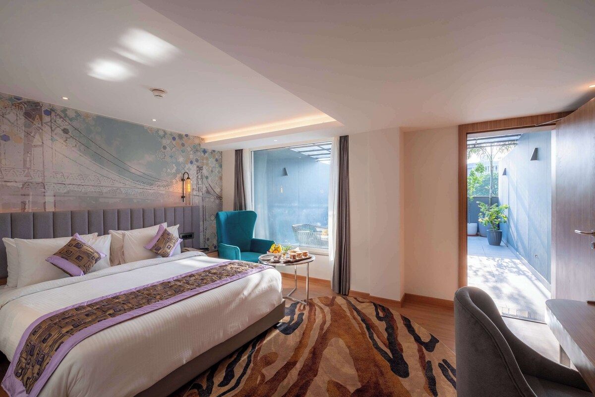 A modern hotel room with a large bed, a blue armchair, a small table with a fruit bowl, and a view of the outdoors through a sliding door.