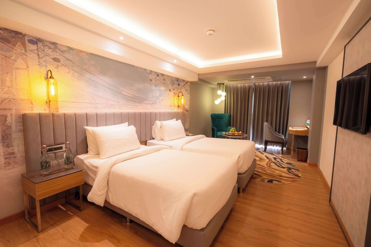 A modern hotel room with two beds, a small table with bottles, a chair, a TV, and a large window with curtains.