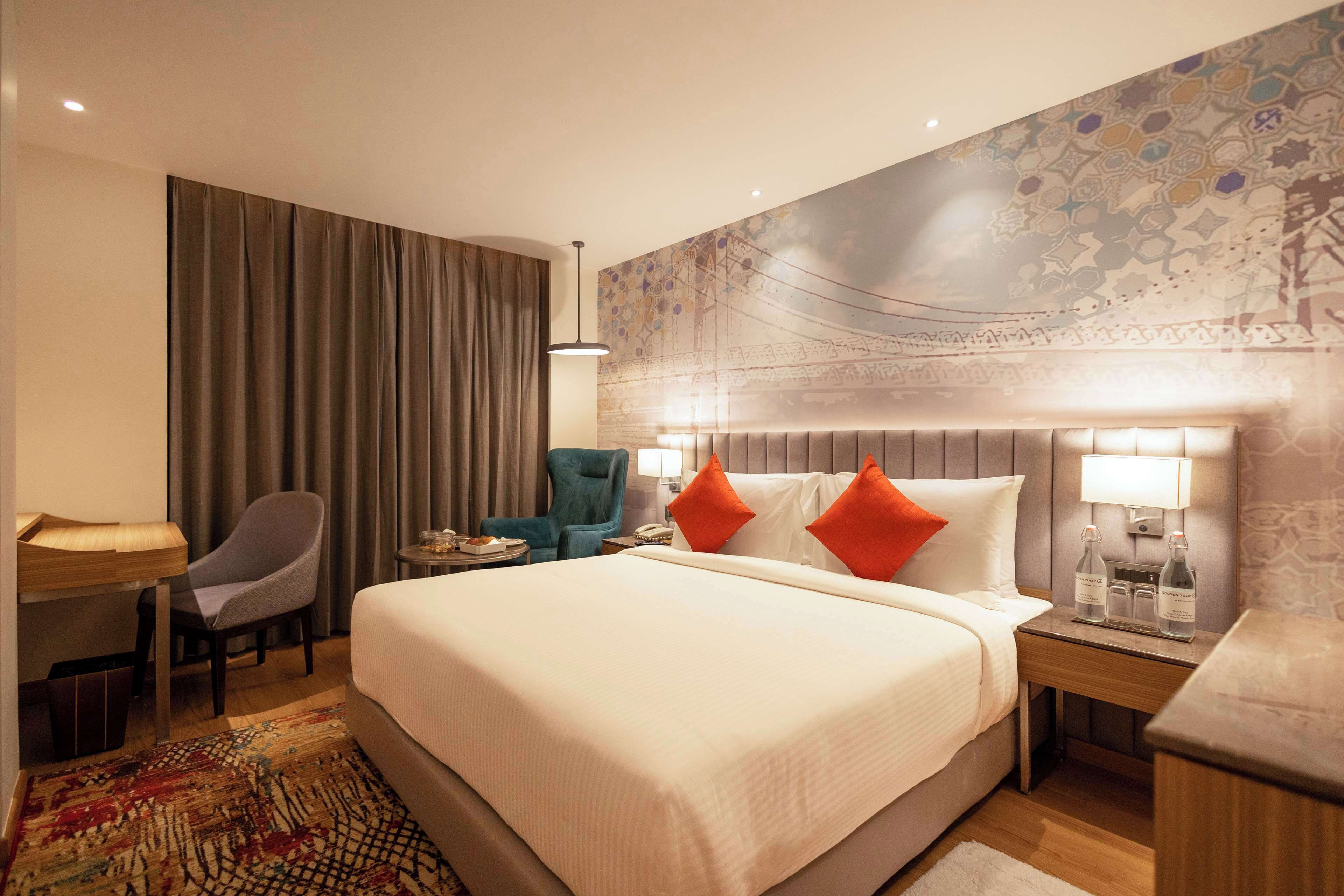 A hotel room with a large bed, a desk, a chair, and a nightstand with a lamp.