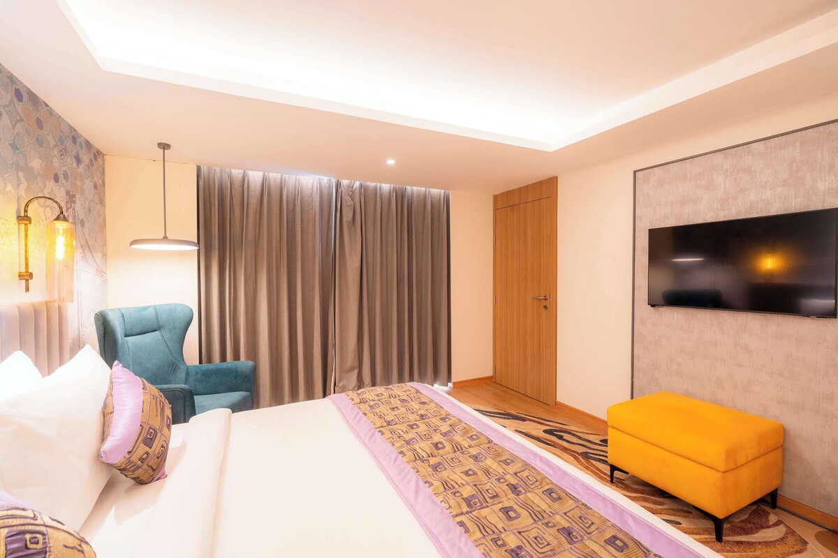 A hotel room with a bed, a blue armchair, a yellow ottoman, a TV mounted on the wall, and a large window with curtains.