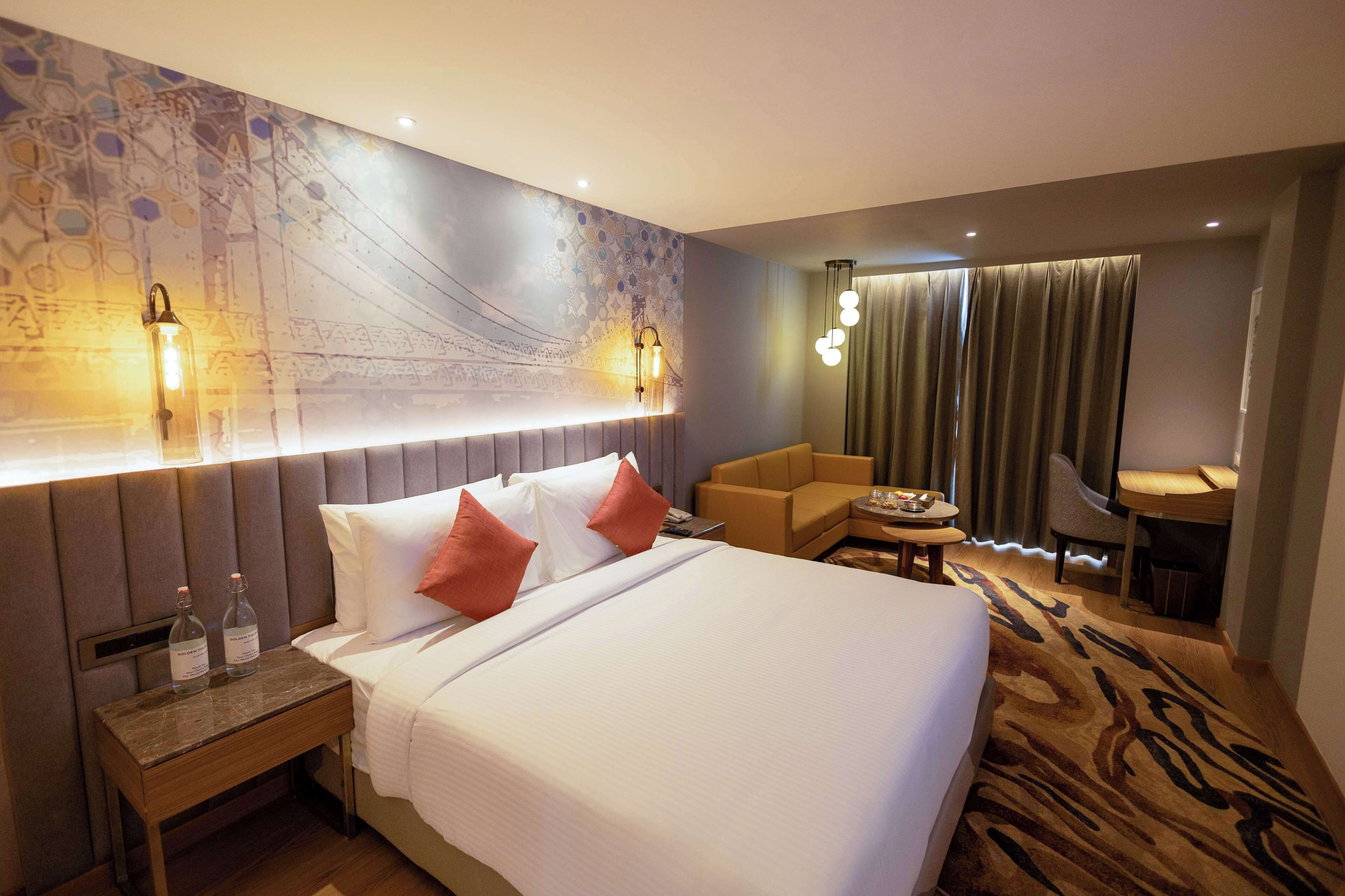 A hotel room with a large bed, a nightstand with two bottles, a sofa, a small table, and a desk.