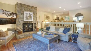 A cozy living room with a stone fireplace, a sofa set, and a dining area with a white railing. - Chetola Resort, Pine #3, in Blowing Rock (Blowing Rock)