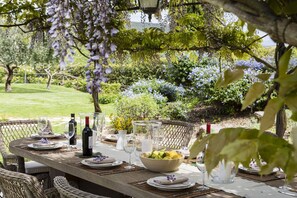 Un área de comedor al aire libre con una larga mesa dispuesta para una comida, rodeada de vegetación exuberante y plantas con flores.