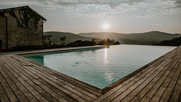 Una piscina infinita con cubierta de madera, rodeada de muros de piedra y vistas a colinas distantes.