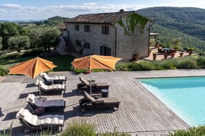 Un espace aménagé au bord de la piscine, avec des chaises longues et des parasols, un bâtiment en pierre et une chaîne de montagnes en arrière-plan.